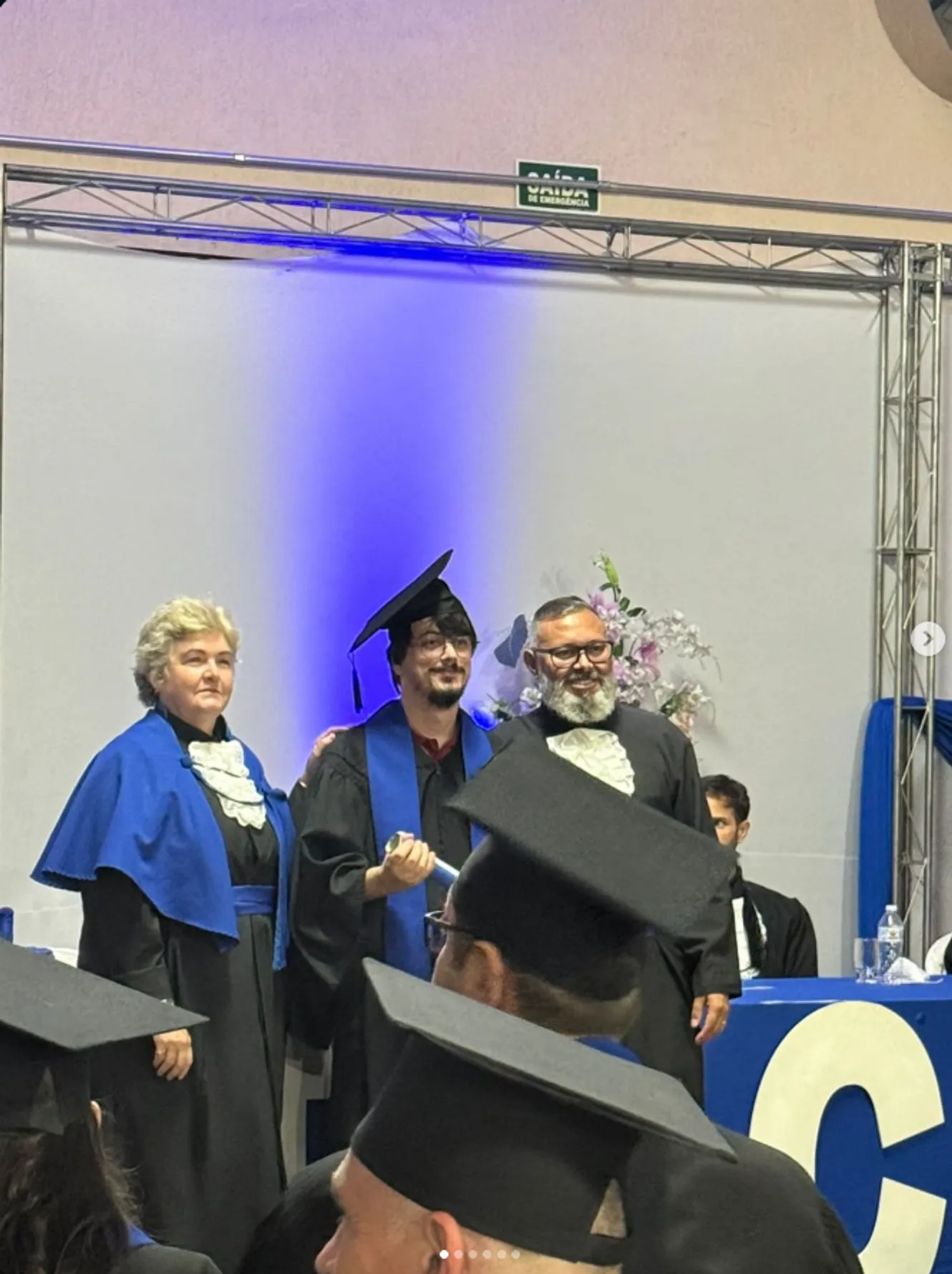 Fotografia original ampliada de cerimônia de formatura, com formando de beca e capelo acompanhado no palco.