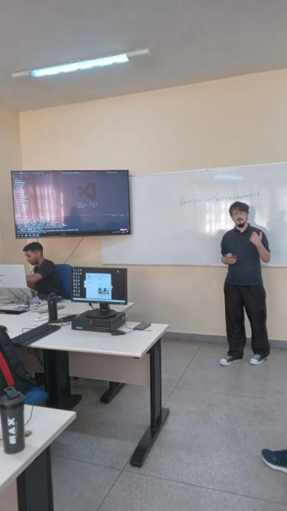 Fotografia original ampliada de apresentação em sala de aula, com código projetado e computadores nas mesas.